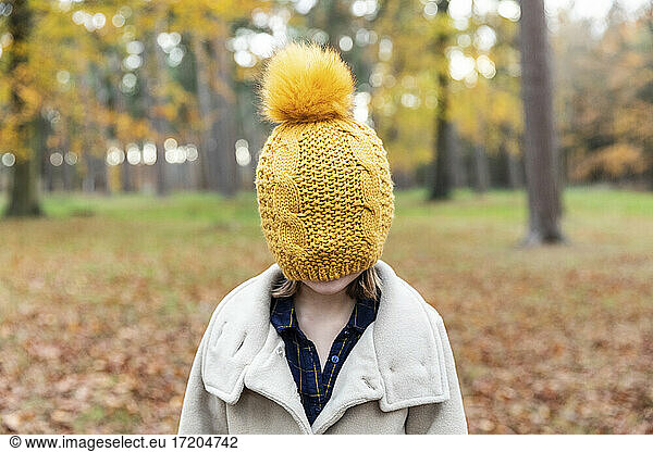 Girl wearing jacket covering face with knit hat while standing at forest
