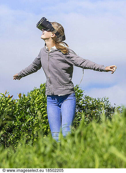 Girl wearing virtual reality headset with arms outstretched on grass field
