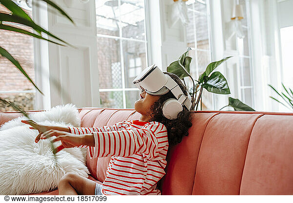 Girl wearing virtual reality simulator gesturing and sitting on sofa at home