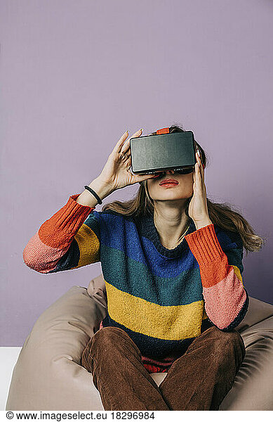 Girl wearing visual reality simulator sitting on bean bag against purple background