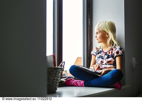 Girl with digital tablet looking through window while sitting on sill