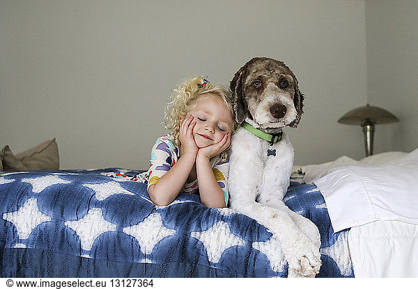 Girl with eyes closed and hands on chin lying by dog on bed against wall
