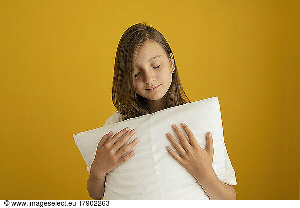Girl with eyes closed holding white pillow against yellow background