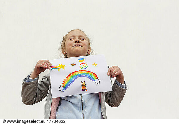 Girl with eyes closed showing rainbow and peace symbol drawing on paper against white background