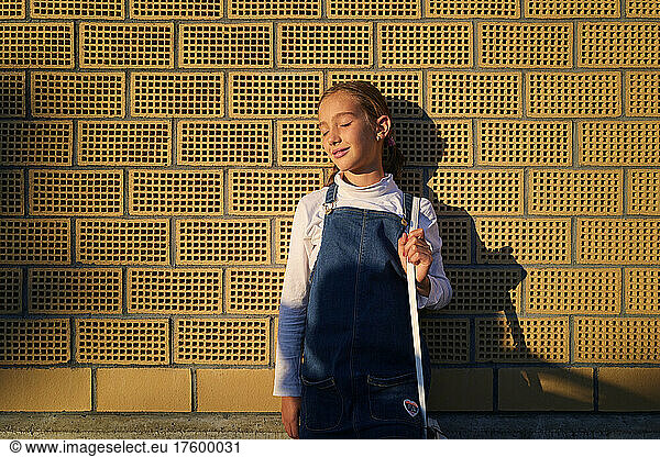 Girl with eyes closed standing in front of brick wall at sunset