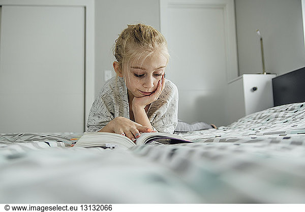 Girl with hand on chin reading book while lying on bed at home