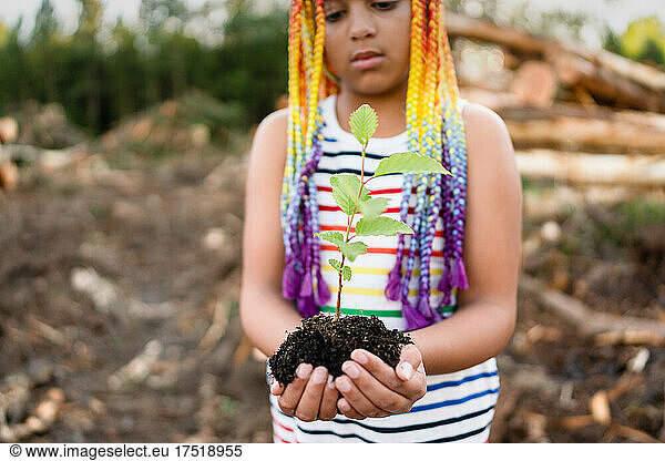 Girl with rainbow braids cups sapling in hands