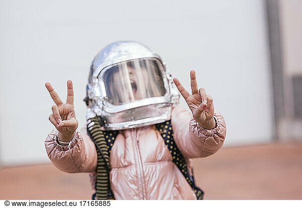 Girl with space helmet gesturing peace sign against white wall