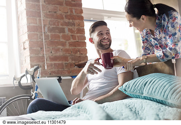Girlfriend bringing coffee to boyfriend using laptop in bedroom