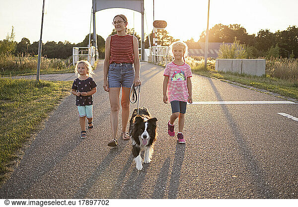 Girls and mother walking with dog on road at sunset