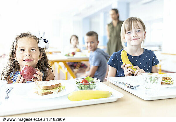 Girls holding apple and banana sitting at table in cafeteria