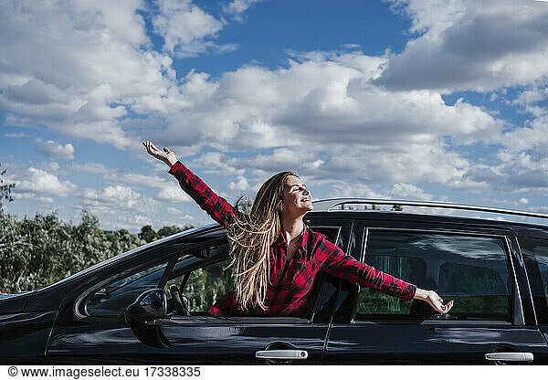 Glückliche blonde Frau mit ausgestreckten Armen  die sich an einem sonnigen Tag aus dem Autofenster lehnt