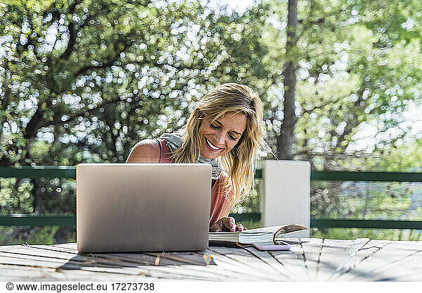Glückliche blonde Touristin sitzt mit Tagebuch und Laptop am Tisch in einem Kurort an einem sonnigen Tag
