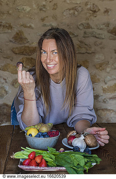 Glückliche Diätassistentin mit verschiedenen Obst- und Gemüsesorten an einem Tisch vor einer Steinwand sitzend