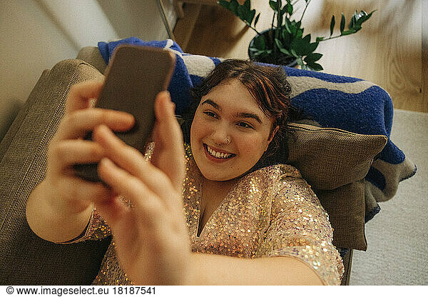 Glückliche Frau  die ein Selfie mit ihrem Smartphone macht  während sie zu Hause auf dem Sofa im Wohnzimmer liegt