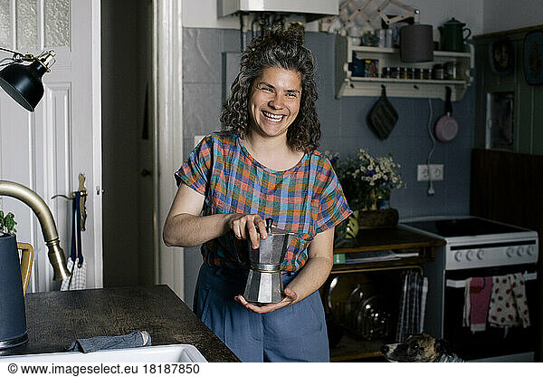 Glückliche Frau  die eine Kaffeekanne in der Hand hält  während sie in der Küche zu Hause steht