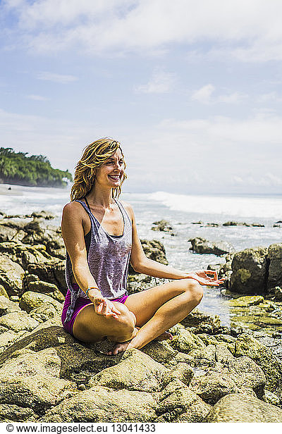 Glückliche Frau im mittleren Erwachsenenalter sitzt im Lotussitz auf Felsen am Strand