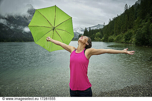 Glückliche Frau mit Regenschirm und ausgebreiteten Armen am Ufer des Haldensees während der Regenzeit
