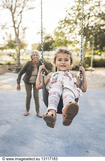 Glückliche Großmutter schiebt Enkelin schaukelnd auf Spielplatz