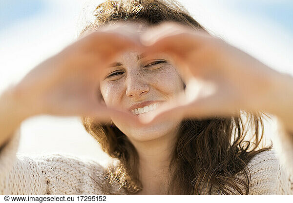 Glückliche junge Frau  die durch ein von Hand geformtes Herz am Strand während eines sonnigen Tages schaut