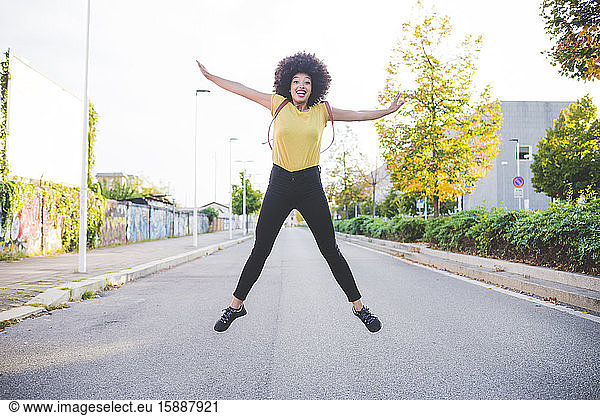Glückliche junge Frau mit Afrofrisur springt auf einer Straße in der Stadt