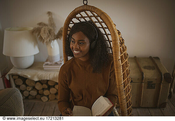 Glückliche junge Frau mit Kopfhörer lesen Buch in hängenden Rattan Stuhl