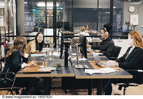 Glückliche junge Geschäftsfrau im Gespräch mit einer Kollegin bei der gemeinsamen Arbeit am Schreibtisch im Büro