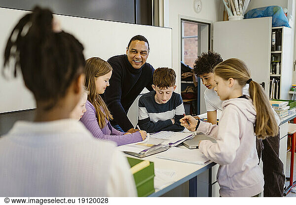 Glückliche Lehrerin mit multirassischen Schülern am Schreibtisch im Klassenzimmer
