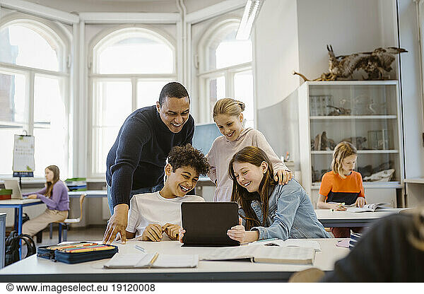 Glückliche männliche und weibliche Schüler  die sich beim Betrachten eines Tablet-PCs mit einem Lehrer im Klassenzimmer vergnügen