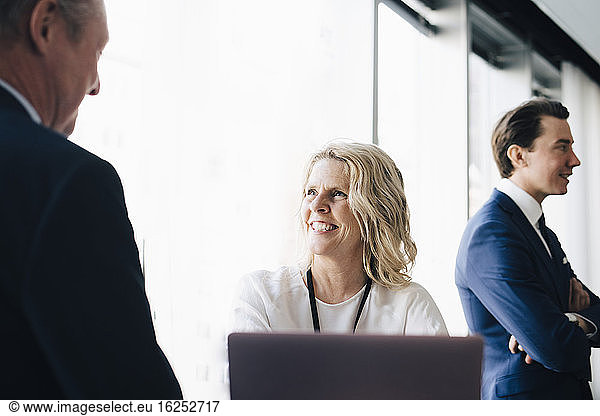 Glückliche Unternehmerin im Gespräch mit einer Kollegin  während die Mitarbeiterin am Arbeitsplatz im Hintergrund steht