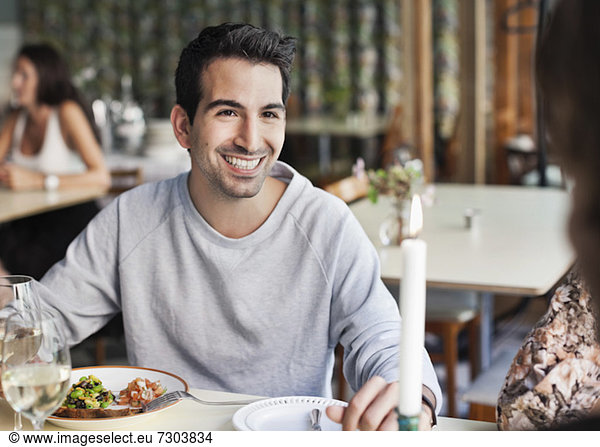 Glücklicher Mann  der eine Freundin am Restauranttisch mit Menschen im Hintergrund ansieht.