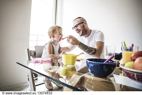 Glücklicher Vater füttert Tochter am Frühstückstisch vor hell erleuchteter Wand