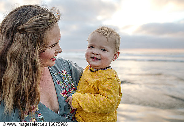 Glowing 30 yr old mom holds happy 6 mo old baby at ocean at sunset