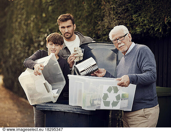 Grandfather  father and son standing by waste bin with recycling boxes with separated waste