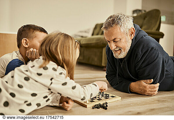 Grandfather looking at girl playing chess lying on floor at home