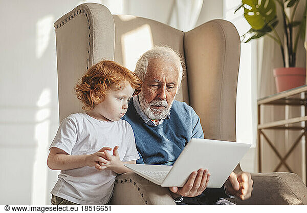 Grandfather sharing laptop sitting in armchair at home