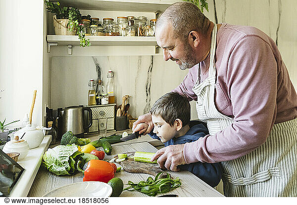 Grandfather teaching grandson to cut vegetables in kitchen at home