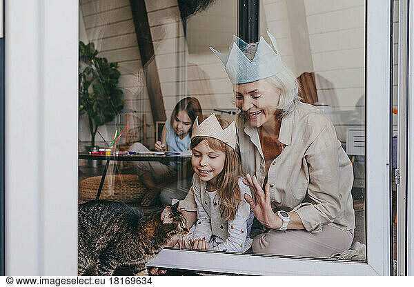 Grandmother and granddaughter wearing crown looking at cat through glass window