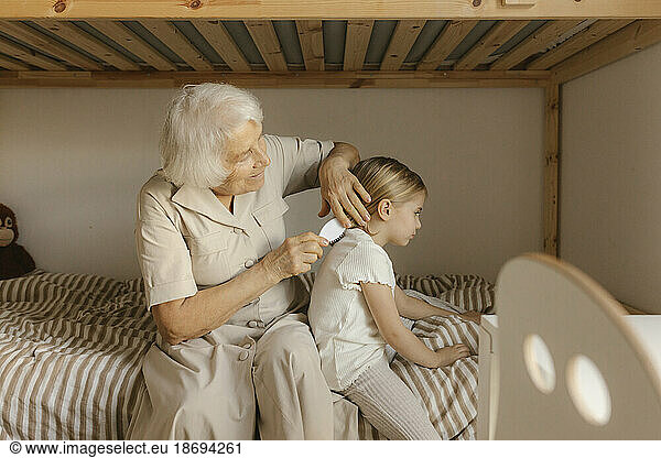 Grandmother combing hair of granddaughter sitting on bed at home