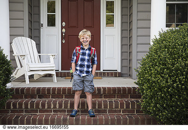 Grinning Blonde Boy With Hands In Pockets Stands on Brick Front Steps