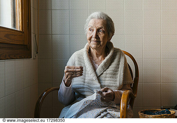 Großmutter näht mit Nadel und Faden  während sie aus dem Fenster schaut