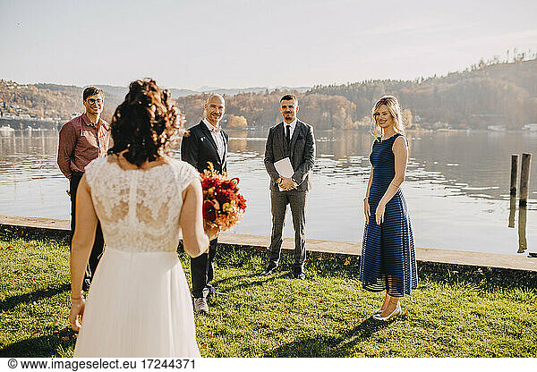 Groom standing with wedding guests looking at bride in front of lake on wedding ceremony