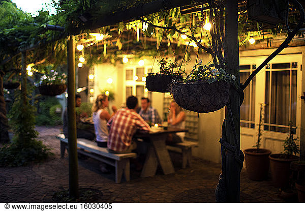 Group of people sitting outdoors at a table  hanging baskets in foreground  evening.