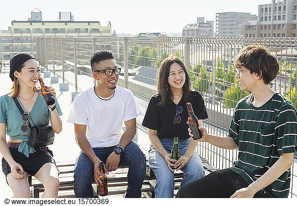 Group of young Japanese men and women sitting on a rooftop in an urban setting  drinking beer.