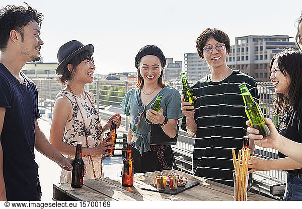 Group of young Japanese men and women standing on a rooftop in an urban setting  drinking beer.