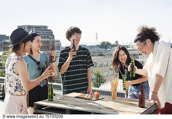 Group of young Japanese men and women standing on a rooftop in an urban setting  drinking beer.