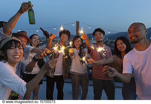 Group of young Japanese men and women with sparklers on a rooftop in an urban setting.