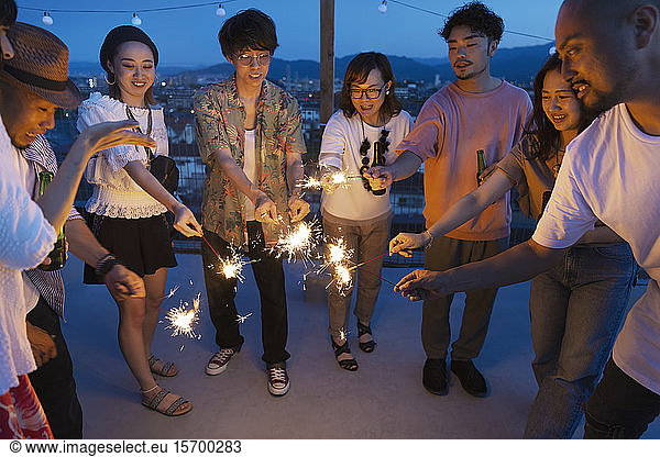 Group of young Japanese men and women with sparklers on a rooftop in an urban setting.