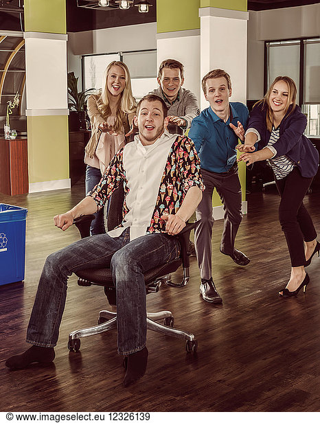 Gruppe junger Millennials  die einen Mitarbeiter in seinem Stuhl quer durch den Raum schieben; Sherwood Park  Alberta  Kanada