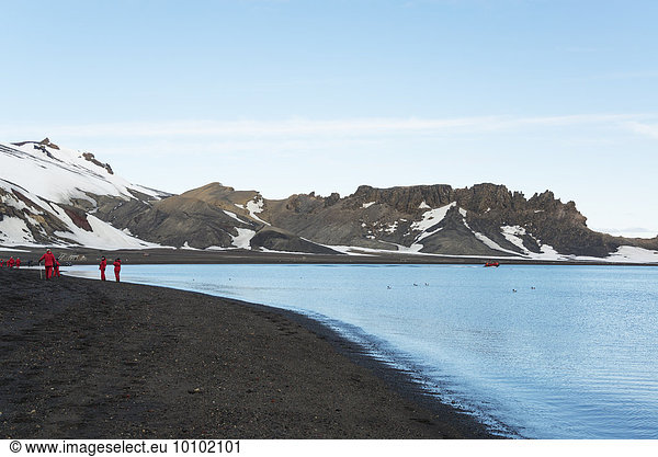 Gruppe von Menschen an einem Strand auf Deception Island  im Hintergrund schneebedeckte Berge.
